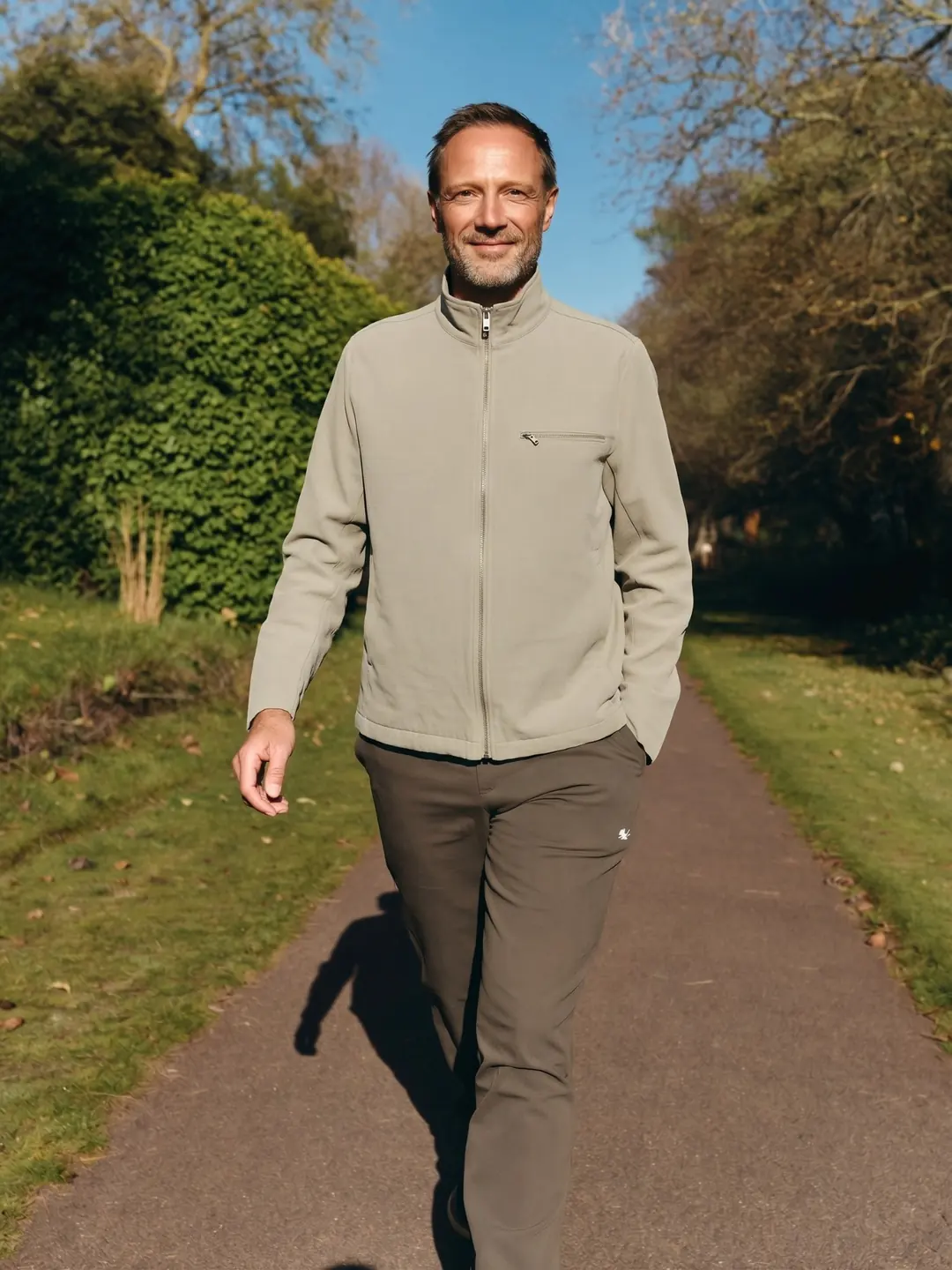 A man in a light zip jacket smiles as he walks along a sunny park path