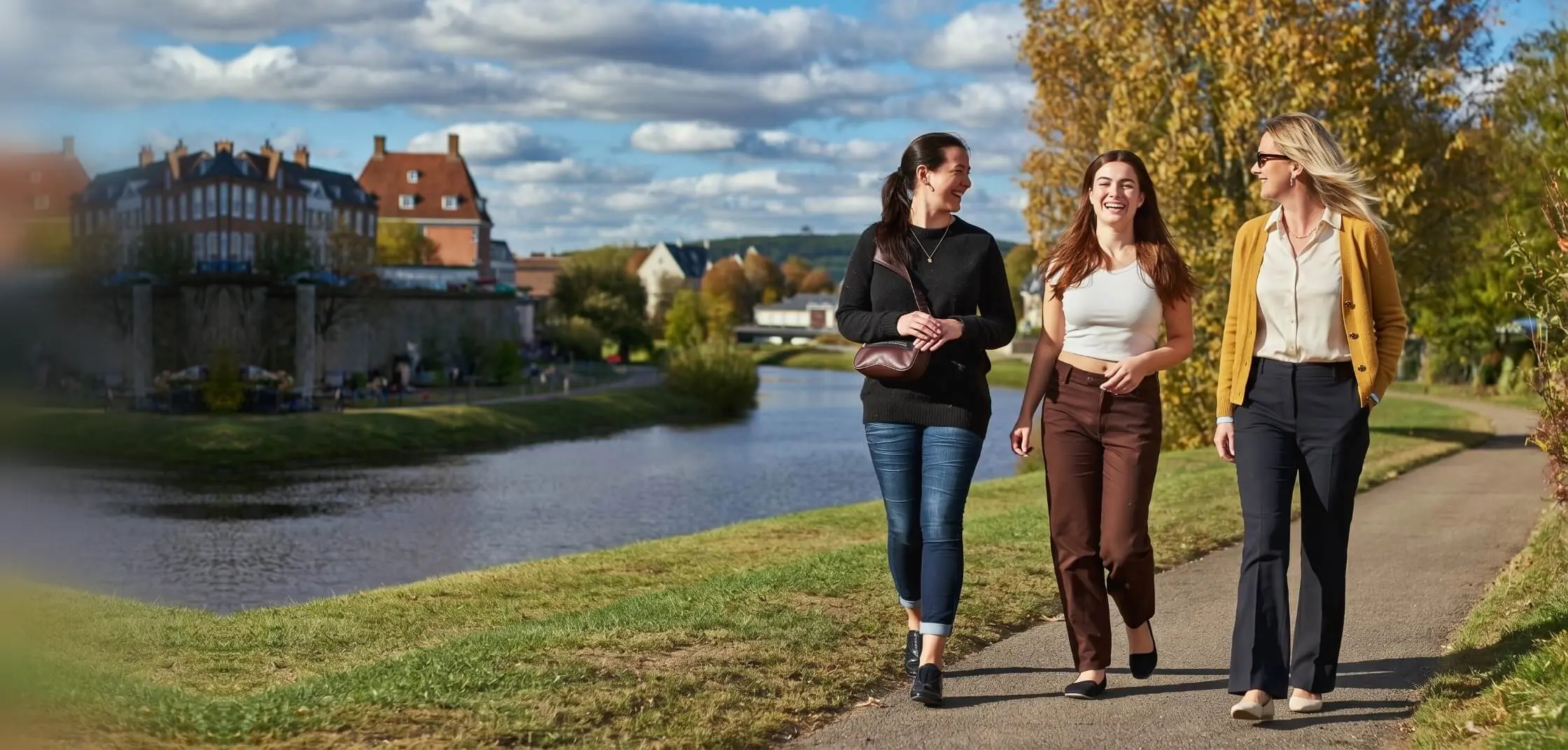 Three women walk and laugh along a riverside path in autumn