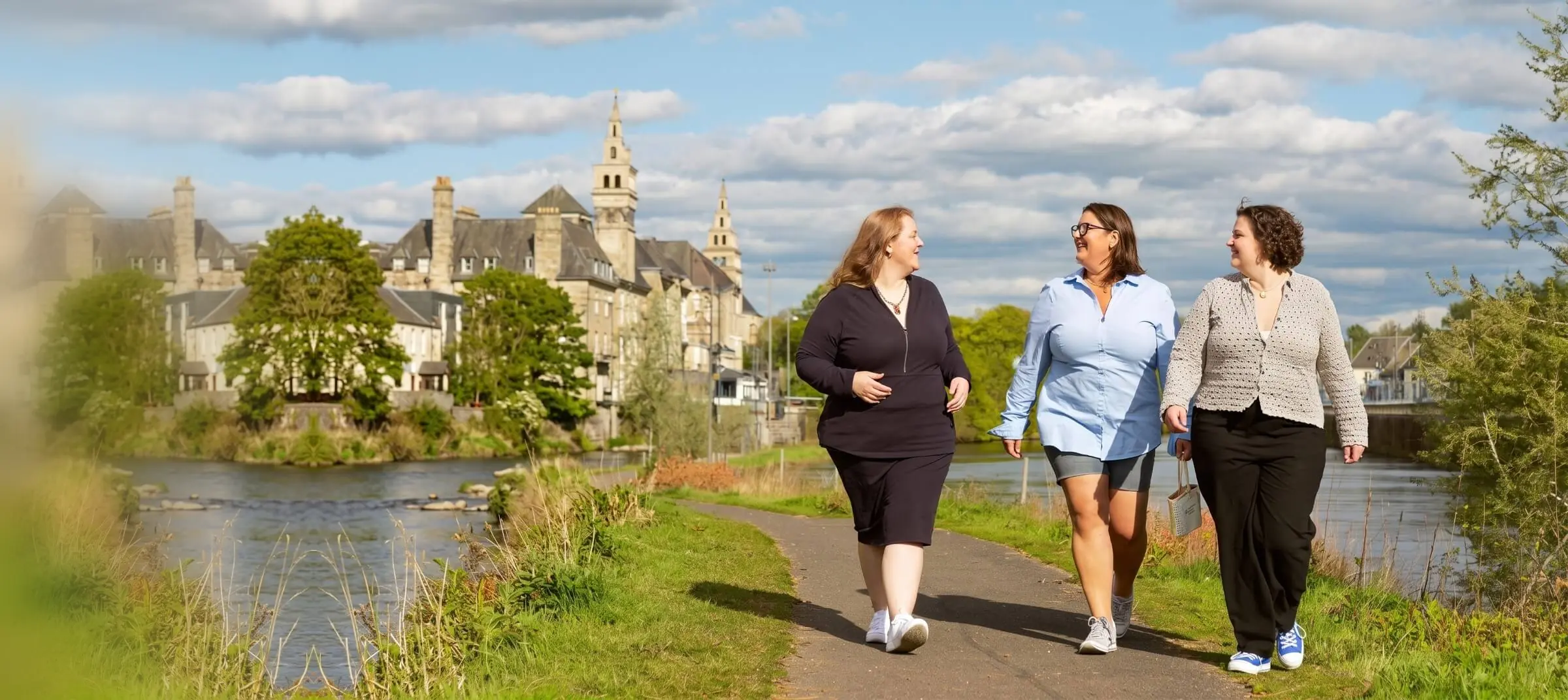 Three women walk and talk along a riverside path on a sunny day