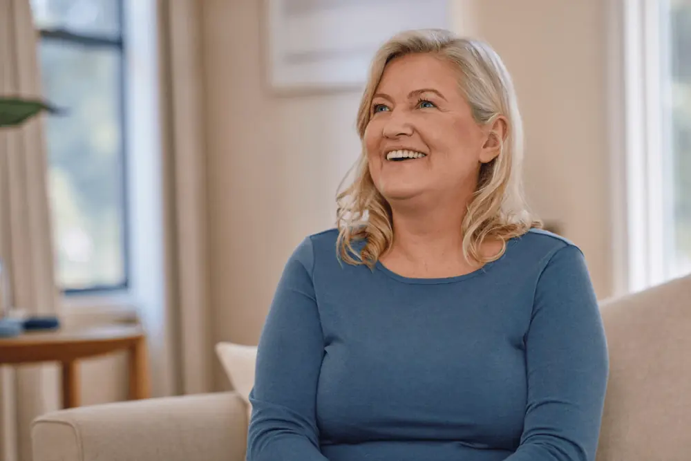 A woman in her fifties smiles on a sofa at home